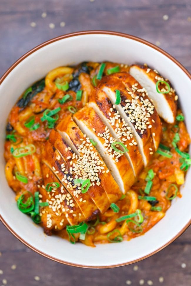 Sliced bulgogi chicken on top of Creamy Dreamy Udon Noodles in bowl on wooden table background with scattered toasted sesame seeds and garnished with spring onion greens.