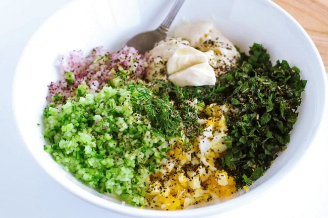 Herby egg salad ingredients unmixed together in large white bowl wit a silver spoon - minced red onion, mayonnaise, minced cucumber, chopped boiled egg, chopped mint leaves, chopped dill leaves, black pepper, kosher salt, and lemon juice. 