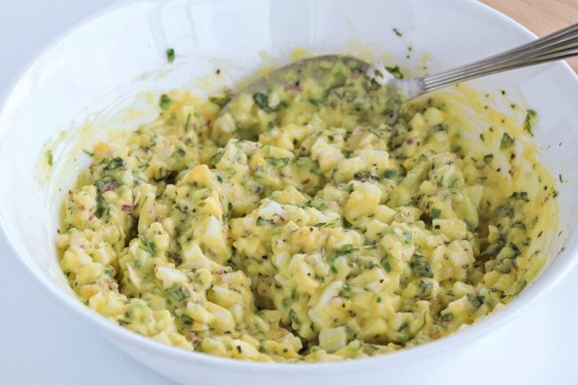Herby egg salad ingredients mixed together in a white bowl with a large silver spoon.