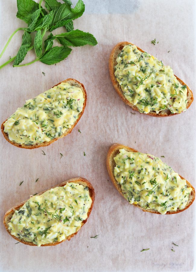 Four herby egg salad tartines sprinkled with fresh dill leaves on top of a nonstick cooking paper background. Mint leaves on stem behind the egg salad tartines - view from top.