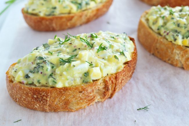 Herby egg salad tartine on a white nonstick cooking paper background with two other tartines cut off in the frame