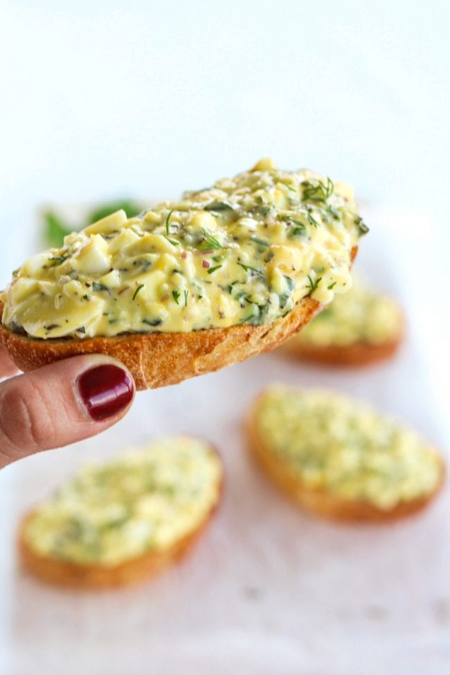 Hand holding up a herby egg salad tartine with three other herby egg salad tartines below on a nonstick cooking paper background
