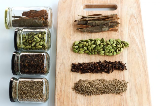 Garam masala ingredients on a chopping board - cassia bark, cardamom pods, whole cloves, cumin seeds. Next to the wooden chopping board are glass airtight jars laid down with each of the ingredients.