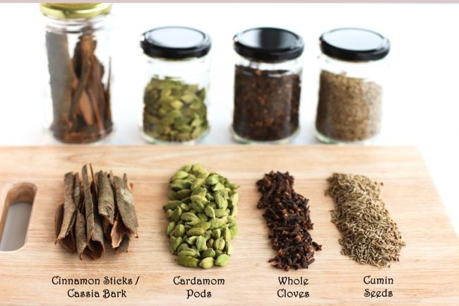 Garam masala ingredients on a chopping board - cassia bark, cardamom pods, whole cloves, cumin seeds. Behind the wooden chopping board are glass airtight jars with each of the ingredients.