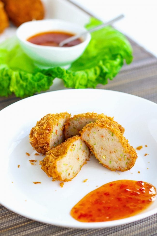 Two spicy Thai shrimp cakes on a white plate cut in half. In the back and diagonally placed to the left is a serving plate with the shrimp cakes, some fresh green lettuce leaves, as well a small serving bowl with Thai sweet chili sauce with a small spoon sticking out of it.