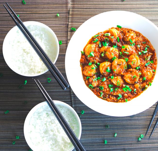 Chili Garlic Shrimp garnished with freshly chopped spring onion greens in a deep white plate on top of a wooden mat background. To the left side of the plate are two bowls of steamed white rice with black chopsticks placed diagonally across them.