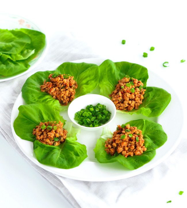 Four Chili Chicken Lettuce Cups on a round white plate placed on top of and gray flower patterned napkin. Small plate of spring onion greens in the center of the plate for garnishing, and plate of butter lettuce leaves on small plate at the top left of photo. Scattered spring onion greens on the white background in front of plate.