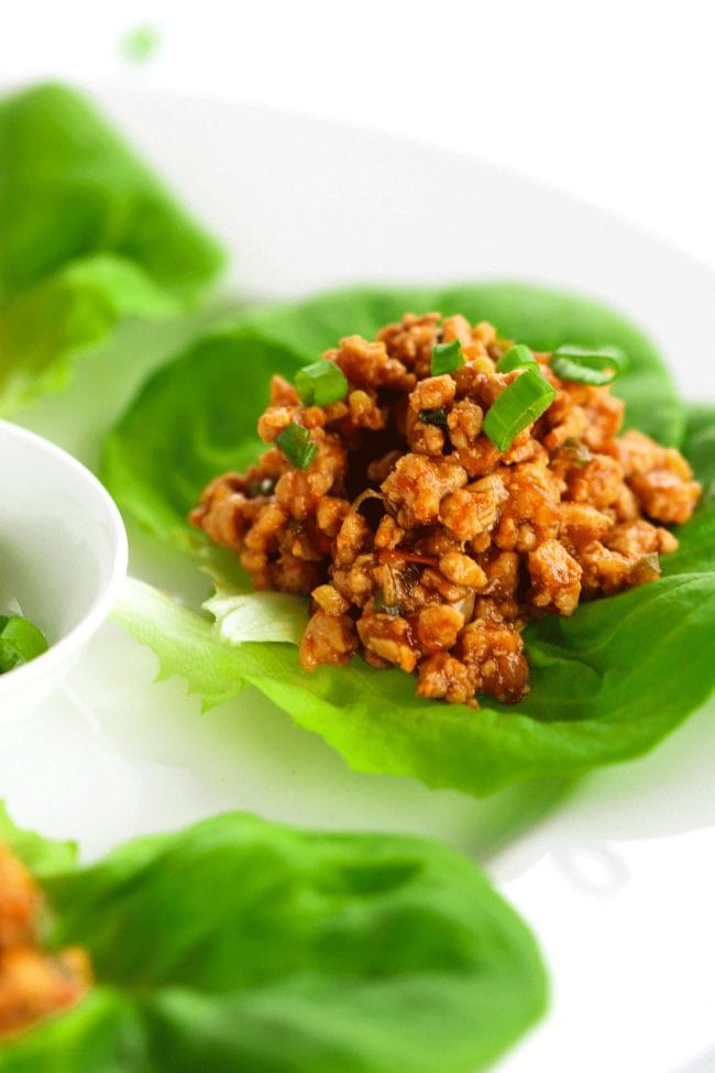 Close up photo of a Chili Chicken Lettuce Cups garnished with spring onion greens on a white round place. In the center of the plate is a small white plate of spring onion greens in the center for garnishing.