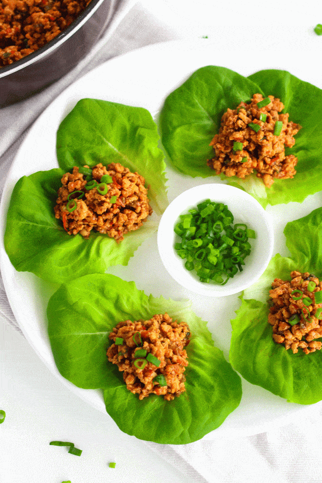 Plate of four chili chicken lettuce cups. In the center of the plate is a small dish of chopped spring onion greens for garnish. To left top hand corner is the sautéed ground chili chicken in a brown colored deep pan. 