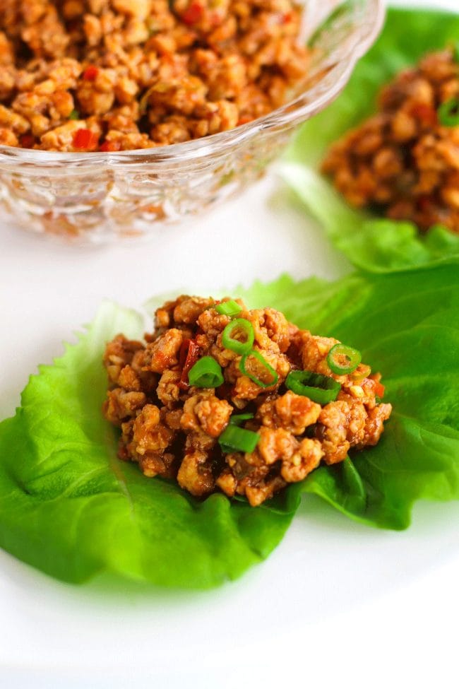 Chili Chicken Lettuce Cups garnished with spring onion greens on a white round place. In the top left of photo (center of plate) is a glass serving bowl filled with chili chicken mince.