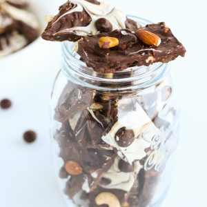 Dark and white chocolate bark pieces with mixed dried fruits and nuts and McVities Dark Chocolate Nibbles stacked in a mason jar on top of a white background. White bowl full of more chocolate bark pieces to left back side of photo.
