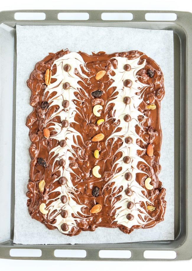 Dark and white chocolate bark (whole unbroken and not yet solid slab) layered with mixed dried fruits and nuts and McVities Dark Chocolate Nibbles layered on top. The chocolate slab is on top of a nonstick cooking paper layered on top of a large baking tray.