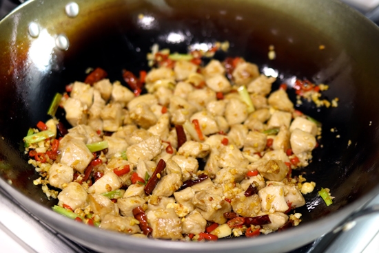 Peanuts, peppercorns, chopped red chilies, minced garlic, minced ginger, spring onion whites, diced chicken pieces and dried red chilies tossed together and cooking in a wok