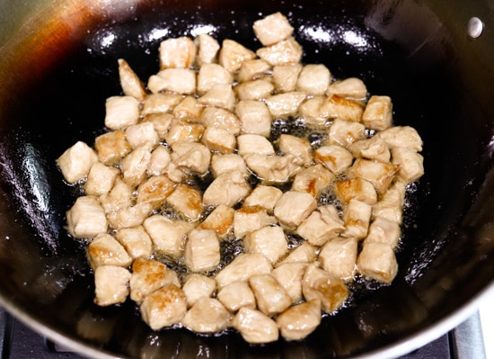 Diced chicken pieces cooking in a wok with peanut oil.