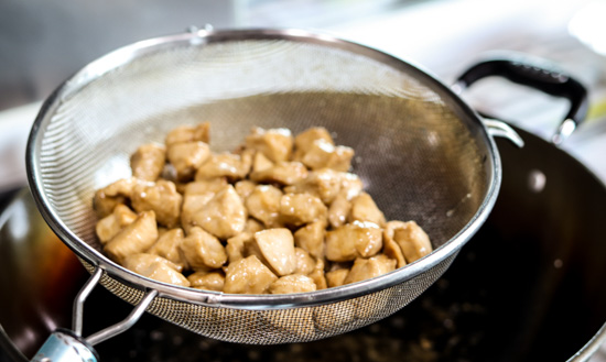 Chicken pieces almost fully cooked in a strainer above a wok.