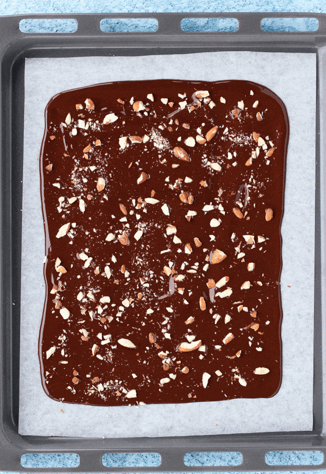 Layer of melted Dark Chocolate with crushed almonds on top of a sheet of nonstick cooking paper on a baking tray on top of a blue bubble patterned backdrop. 