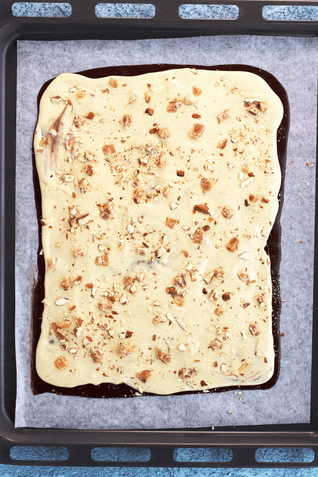 Layer of melted Dark Chocolate with crushed almonds and layer of white chocolate with some more crushed almonds and candied ginger on top of a sheet of nonstick cooking paper on a baking tray on top of a blue bubble patterned backdrop. 