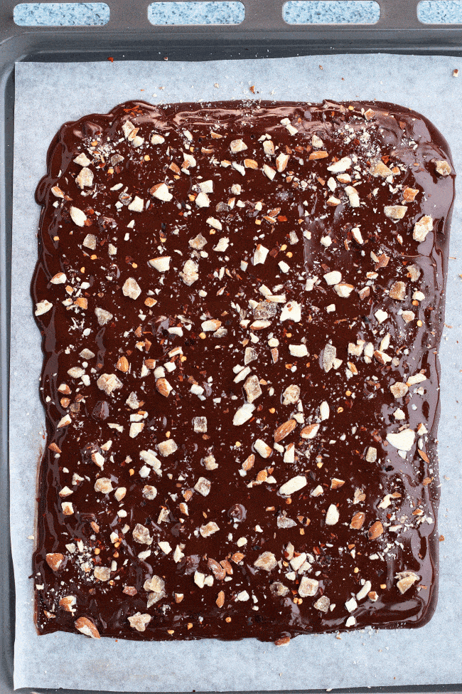 Spicy Chocolate Bark with Almonds & Candied Ginger with final layer of ayer of melted Dark Chocolate with crushed almonds, extra candied ginger, and chili flakes sprinkled on top. Placed on top of a sheet of nonstick cooking paper on a baking tray on top of a blue bubble patterned backdrop. 