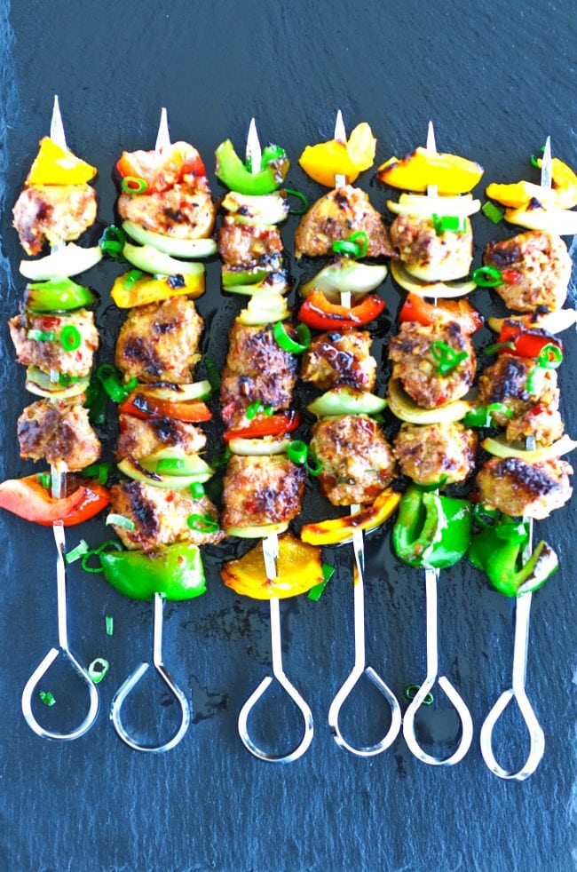Perfectly grilled marinated chicken pieces, diced bell pepper, and diced onion threaded on metal skewers on top of a black stone plate background. Garnished with chopped spring onion greens. 