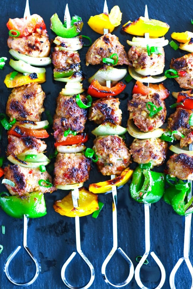 Perfectly grilled marinated chicken pieces, diced bell pepper, and diced onion threaded on metal skewers on top of a black stone plate background. Garnished with spring onion greens. 