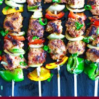 Perfectly grilled marinated chicken pieces, diced bell pepper, and diced onion threaded on metal skewers on top of a black stone plate background. Garnished with spring onion greens.