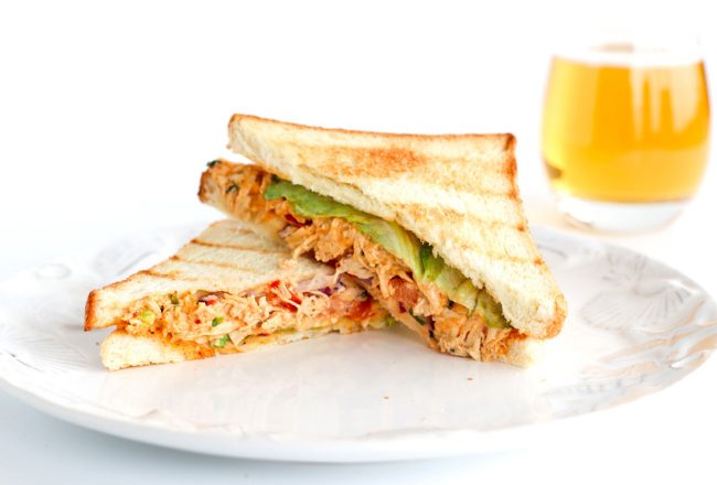 Front view of diagonally sliced Sweet & Spicy Thai Chicken Salad Sandwich stacked at a slanted angle on top of white plate. Glass of apple juice behind the plate.