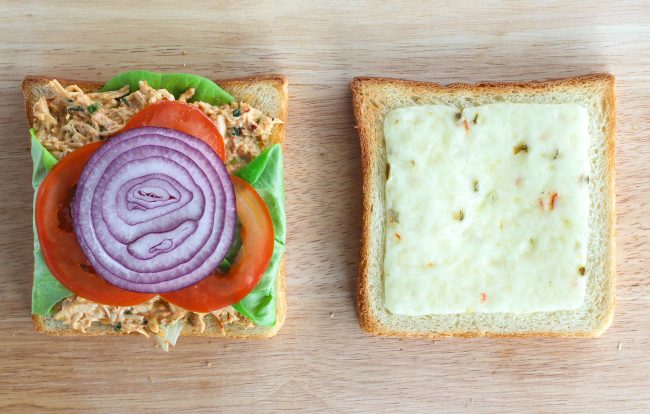 Toasted white bread slices on wooden chopping board - one with  lettuce, Sweet & Spicy Thai Chicken Salad, tomato slices, and red onion on top. The other with pepper jack cheese on top. 