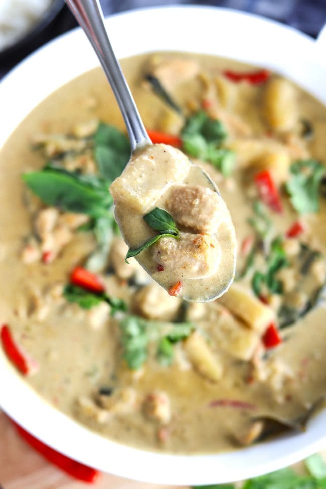 Overhead view of a silver spoon holding up a bite of Thai Green Chicken Curry above a large white serving bowl with the rest of the curry. 