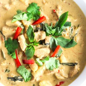 Thai Green Chicken Curry in a large deep white serving bowl on top of a round wooden chopping board. The curry is garnished with coriander, basil leaves, and red chili strips. Bowl is cut off from the photo from the left side.