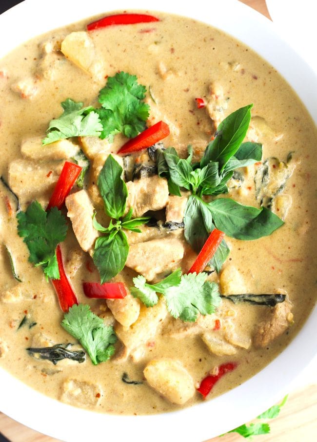 Thai Green Chicken Curry in a large deep white serving bowl on top of a round wooden chopping board. The curry is garnished with coriander, basil leaves, and red chili strips. 