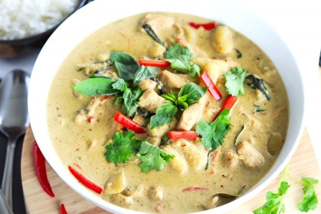 Thai Green Chicken Curry in a large deep white serving bowl on top of a round wooden chopping board. The curry is garnished with coriander, basil leaves, and red chili strips. Bowl of steamed white rice, silver spoon, and white soup ladle to the side. 