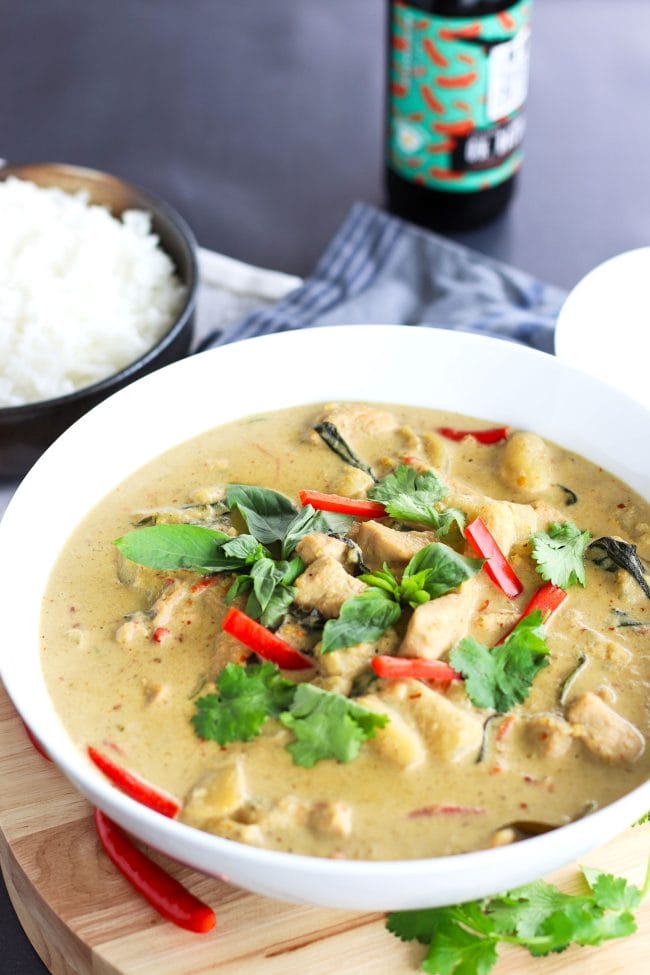 Thai Green Chicken Curry in a large deep white serving bowl on top of a round wooden chopping board which is on top of a blue striped kitchen towel. The curry is garnished with coriander, basil leaves, and red chili strips. Bowl of steamed white rice and white soup ladle to the side. Bottle of ice cold beer in the back on top of a chalkboard backdrop. 