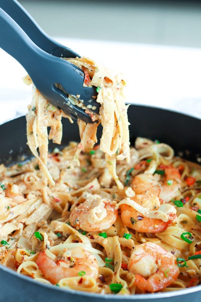 Black pasta tongs holding up a serving of Garlic Cream Sauce Seafood Fettuccine above a black pan of the pasta.