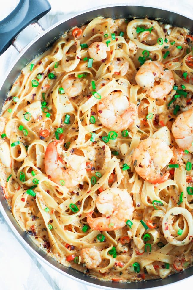 Black pan with Garlic Cream Sauce Seafood Fettuccine cut off from left side of photo. Jumbo juicy shrimp are on top of a bed of pasta in the pan. Garnished with spring onion greens and crushed red pepper flakes.