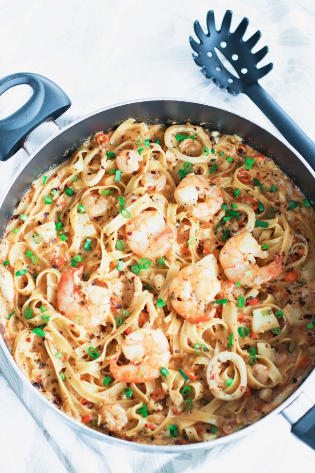 Black pan with Garlic Cream Sauce Seafood Fettuccine cut off from left side of photo. Jumbo juicy shrimp are on top of a bed of pasta in the pan. Garnished with spring onion greens and crushed red pepper flakes. Black pasta server to the side of pan.