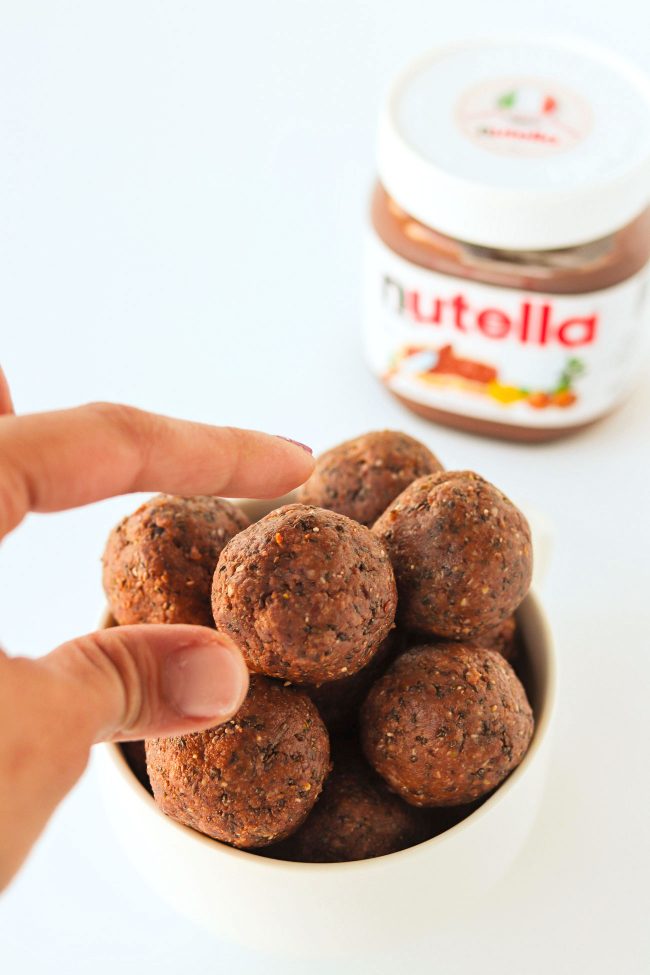 Hand reaching for a Nutella & Peanut Butter Energy Ball from a bowl that is stacked high with energy balls. Jar of Nutella behind the bowl on the right side of the photo.