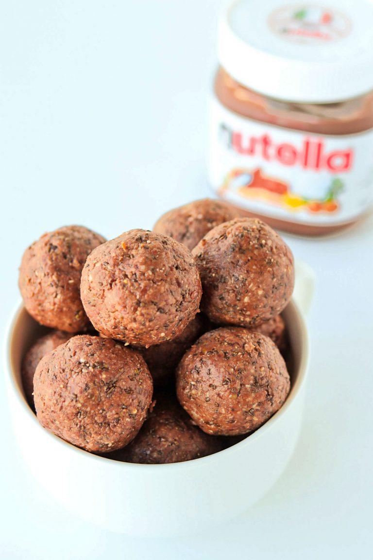 White bowl of Nutella & Peanut Butter Energy Balls stacked high with two balls in front of the bowl on a white backdrop. Jar of Nutella on the back right side of the bowl.