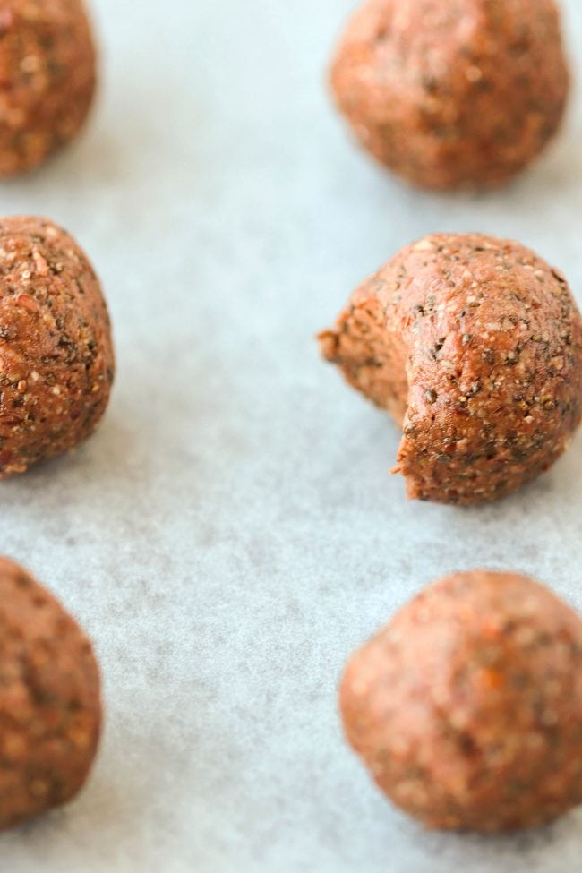 Six Nutella & Peanut Butter Energy Balls lined up on top of non-stick cooking paper. Close up and focus on one energy ball that has a bite taken out of it.