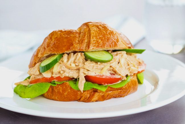 Spicy Asian-Cajun Chicken Salad Croissandwich made with tomatoes, butter lettuce, cucumber slices on top of a white plate on a chalkboard backdrop. Glass of sparkling water in the back.
