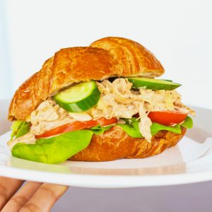 Hand holding up a white plate with a Spicy Asian-Cajun Chicken Salad Croissandwich made with tomatoes, butter lettuce, cucumber slices over a chalkboard backdrop.