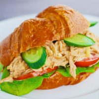 Spicy Asian-Cajun Chicken Salad Croissandwich made with tomatoes, butter lettuce, cucumber slices on top of a white plate on a chalkboard backdrop.
