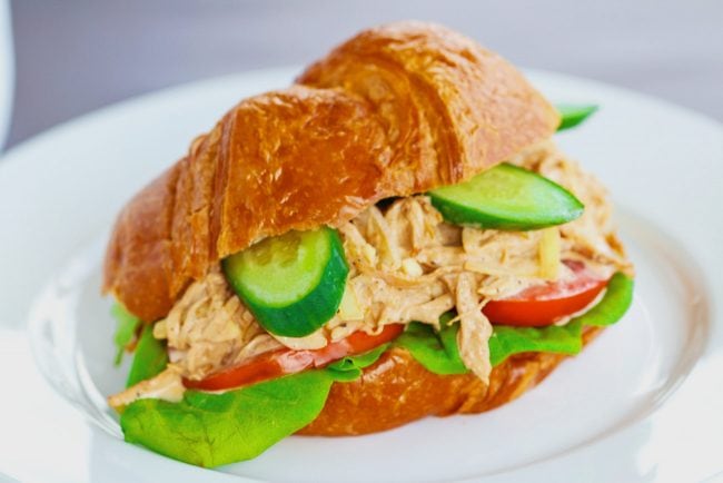 Spicy Asian-Cajun Chicken Salad Croissandwich made with tomatoes, butter lettuce, cucumber slices on top of a white plate on a chalkboard backdrop.