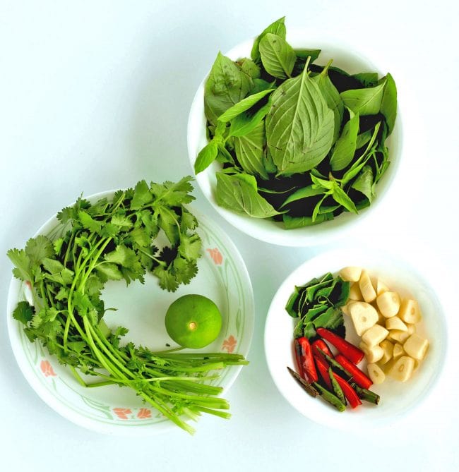 Bowl with Thai Sweet Basil Leaves, bowl with coriander and a lime, and a bowl with torn up kaffir lime leaves, garlic cloves, and roasted halved red and green chilies.