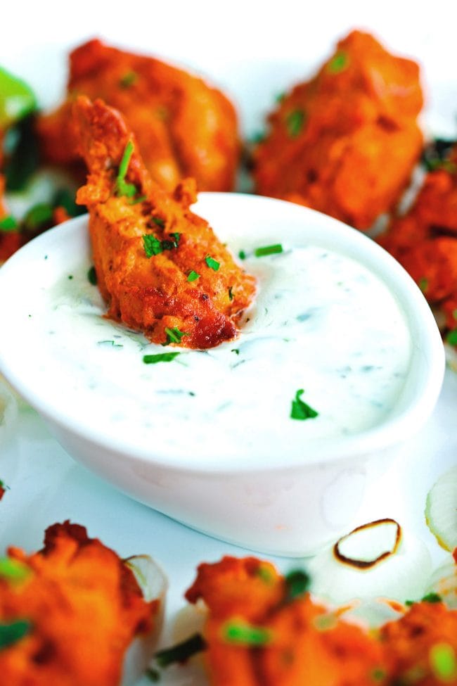 A chicken tikka drumette in a dipped into a small dish of cucumber and mint yogurt dip surrounded by chicken tikka drumettes on a white plate.