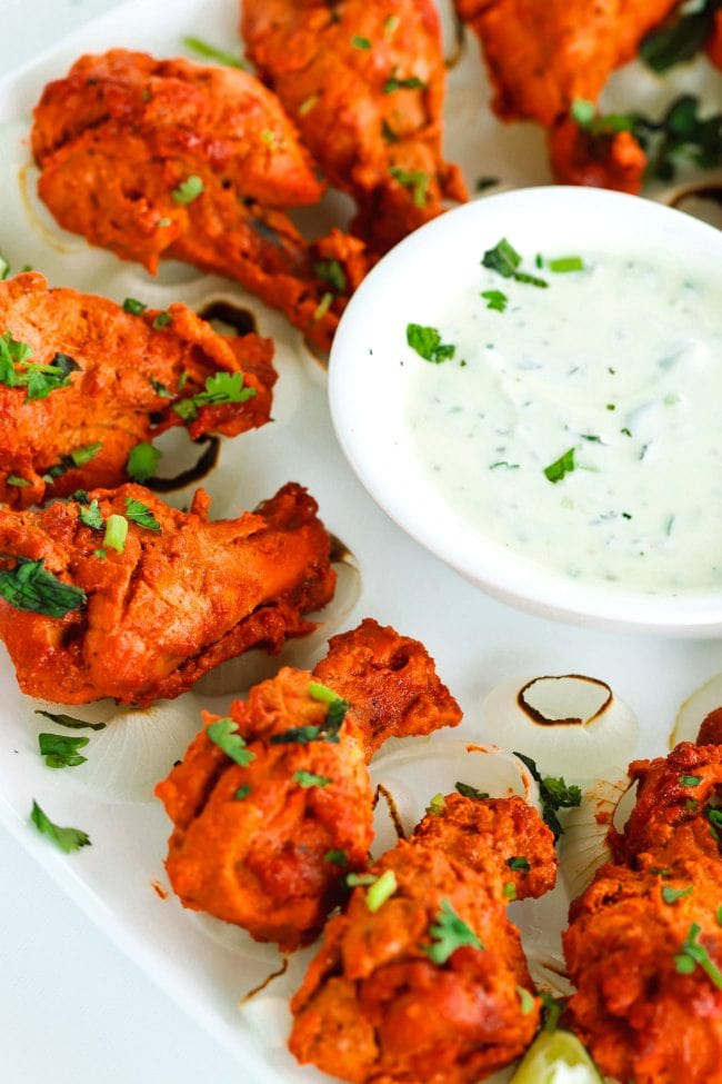 Close up of left side of square plate loaded with oven grilled chicken tikka drumettes, and a small dish of cucumber and mint yogurt dip in the center of the plate.