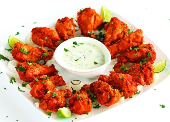 A white square plate of chicken tikka drumettes over a bed of chargrilled onion rings. Garnished with freshly chopped coriander. Lime wedges in the corners of the plate and in the center is a small dipping dish with cucumber and mint yogurt dip.