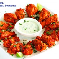 A white square plate of chicken tikka drumettes over a bed of chargrilled onion rings. Garnished with freshly chopped coriander. Lime wedges in the corners of the plate and in the center is a small dipping dish with cucumber and mint yogurt dip.