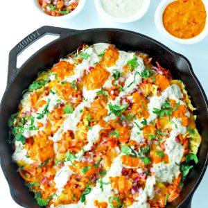 Overhead view of cast iron skillet of Chicken Tikka Nachos with Creamy Cooling Yogurt Sauce and Red Chili sauce drizzled over the top. Garnished with freshly chopped coriander. Above the skillet are three dip bowls filled with pico de gallo, creamy cooling yogurt sauce, and a red chili sauce.