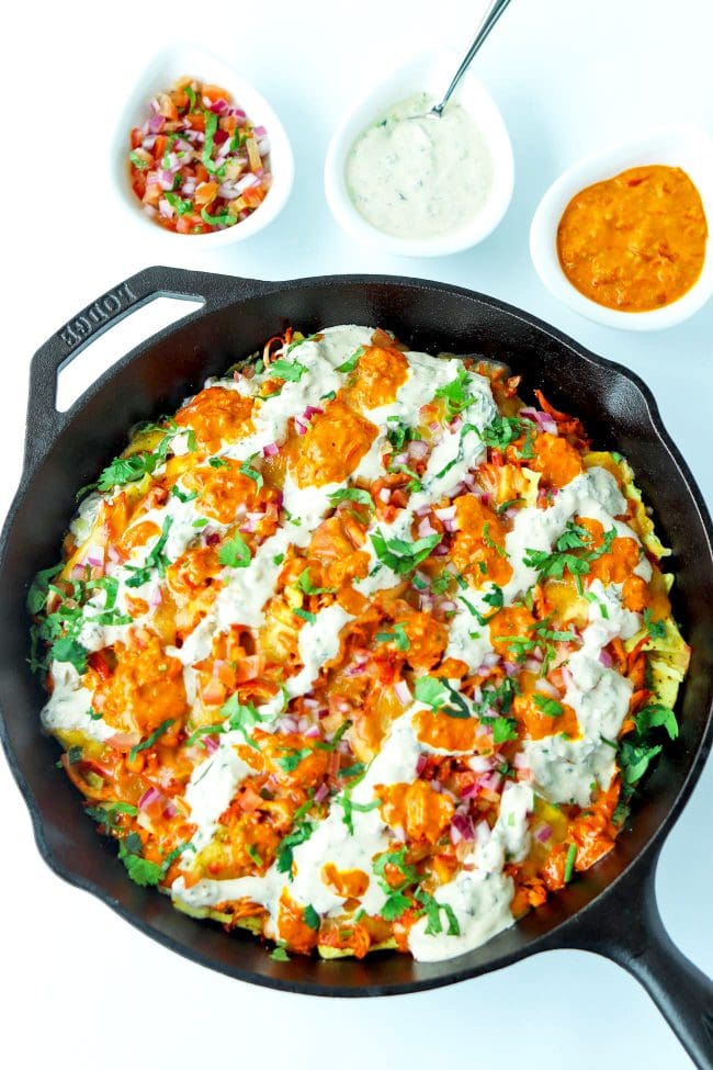 Overhead view of cast iron skillet of Chicken Tikka Nachos with Creamy Cooling Yogurt Sauce and Red Chili sauce drizzled over the top. Garnished with freshly chopped coriander. Above the skillet are three dip bowls filled with pico de gallo, creamy cooling yogurt sauce, and a red chili sauce.