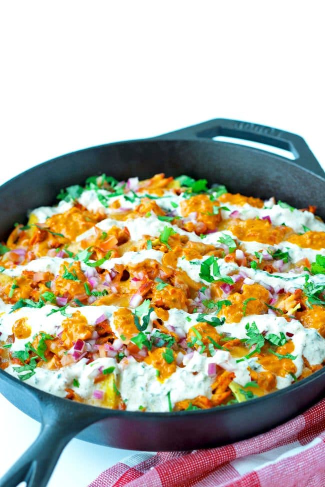 Front side view of cast iron skillet on red and white checkered patterned napkin with Chicken Tikka Nachos, topped with Creamy Cooling Yogurt Sauce and Red Chili sauce drizzled over the top. Garnished with freshly chopped coriander.